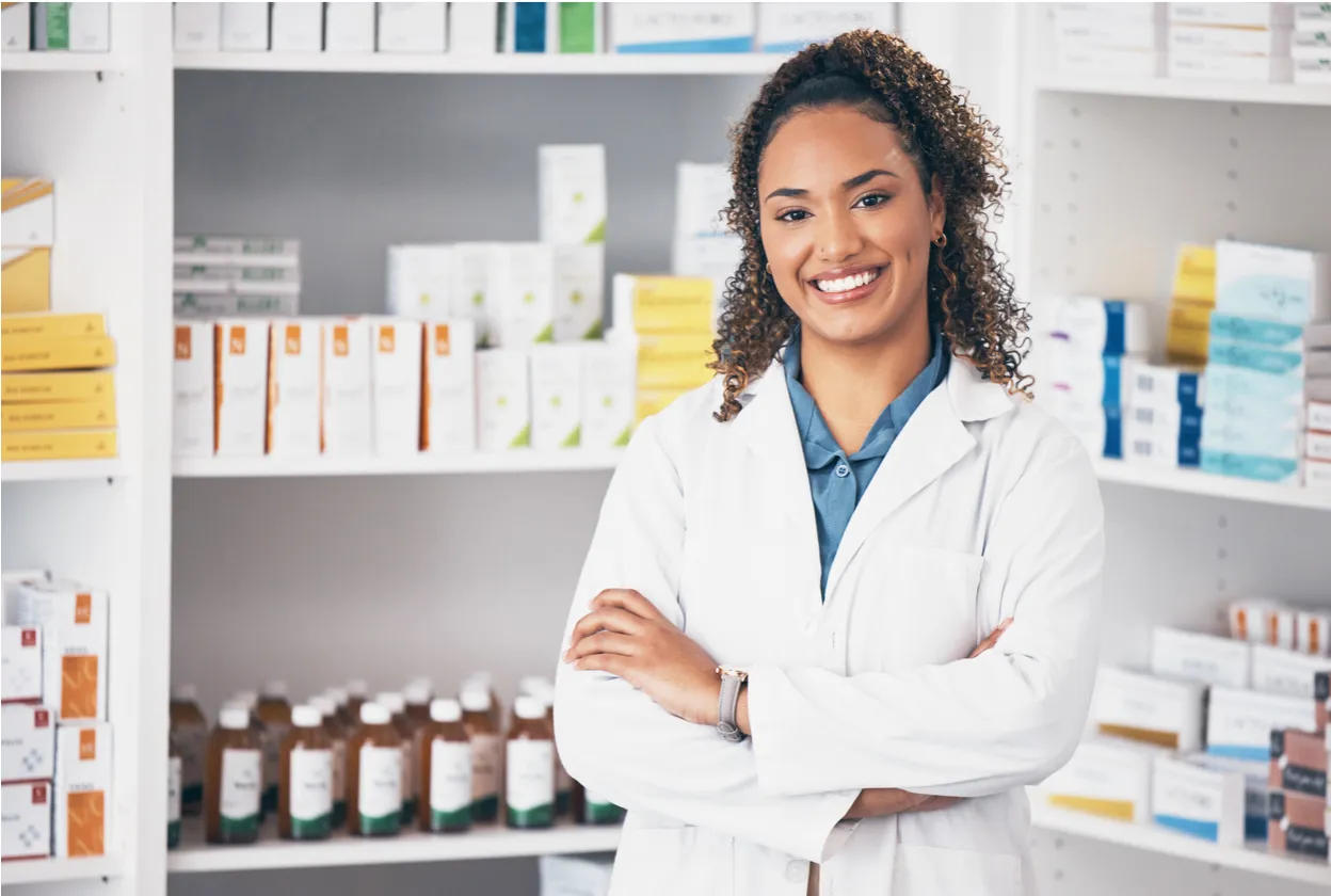 Smiling female pharmacist in white coat standing with arms crossed in front of pharmacy shelves filled with medications and supplies